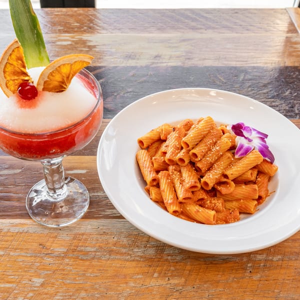 Rigatoni pasta with red sauce and a purple orchid, next to a frozen cocktail with fruit garnishes.