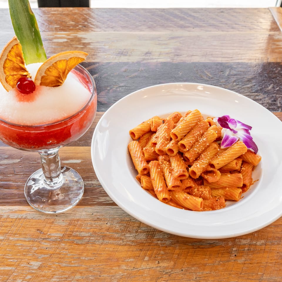 Rigatoni pasta with red sauce and a purple orchid, next to a frozen cocktail with fruit garnishes.