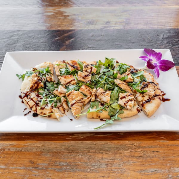 Chicken flatbread with arugula and balsamic glaze, garnished with a purple orchid on a white plate.